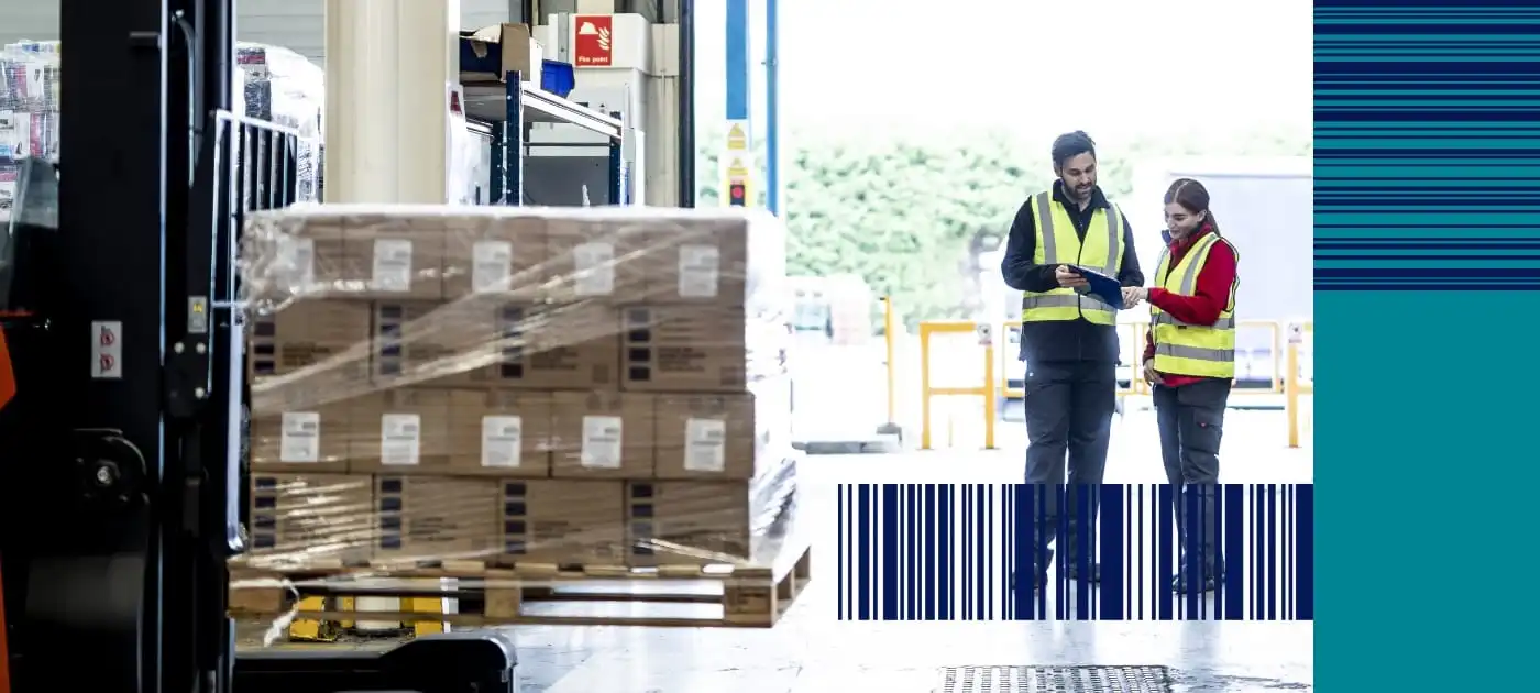 Male and female warehouse workers wearing high visibility vests are looking at a clipboard, a forklift vehicle with a pallet of brown cardboard boxes is in the foreground
