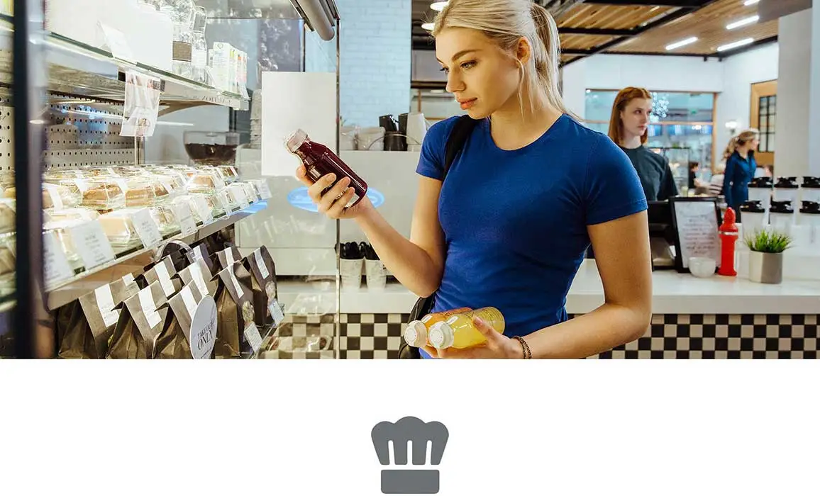 Woman with blond hair wearing blue tshirt looking at a juice bottle holding sandwich
